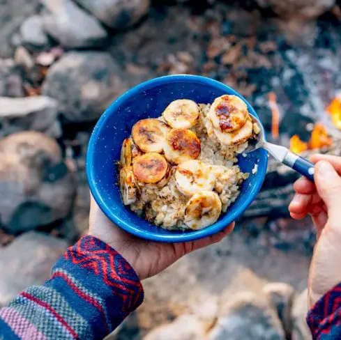 Megan holding a blue bowl of oatmeal topped with bananas and campfire in the background
