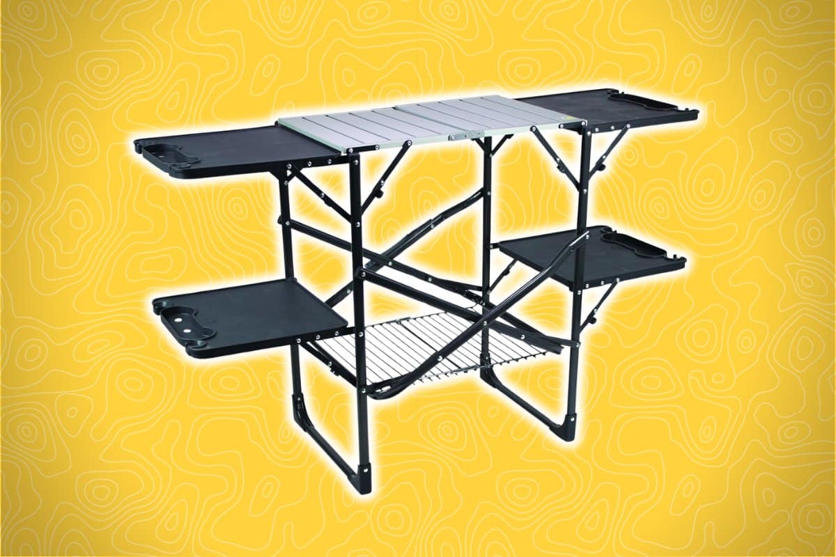 A collapsible camping kitchen table with multiple shelves, a metal tabletop, and folding black legs is set against a yellow background.