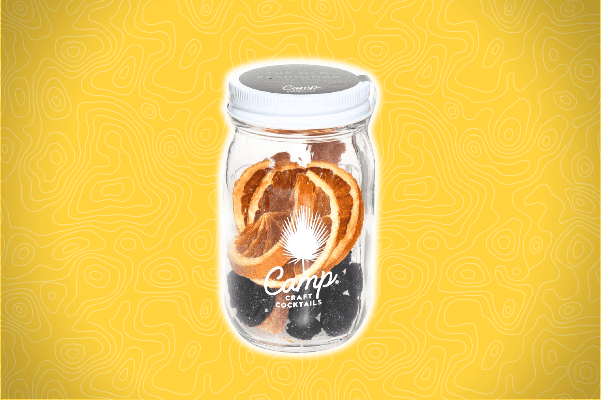 A glass jar labeled "Camp Craft Cocktails" contains dried orange slices and botanicals, set on a bright yellow background.