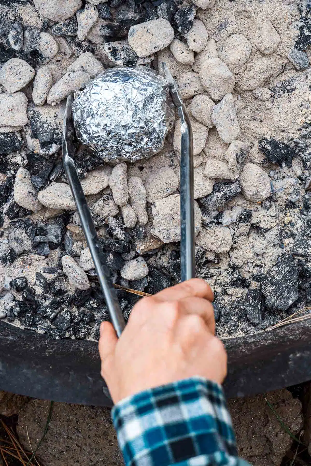 Using tongs to place a foil wrapped apple into a campfire.