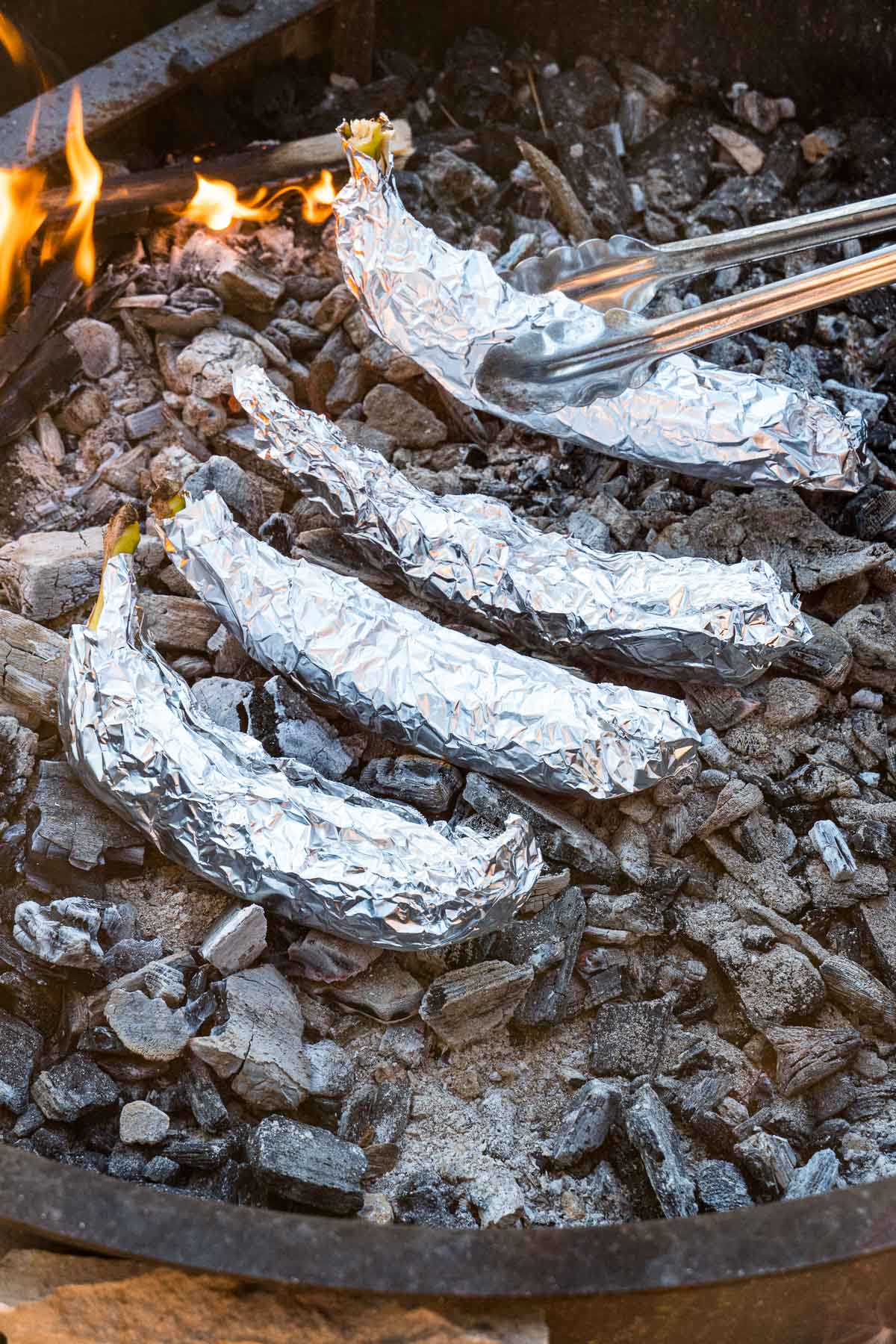 Four bananas wrapped in foil in the embers of a campfire