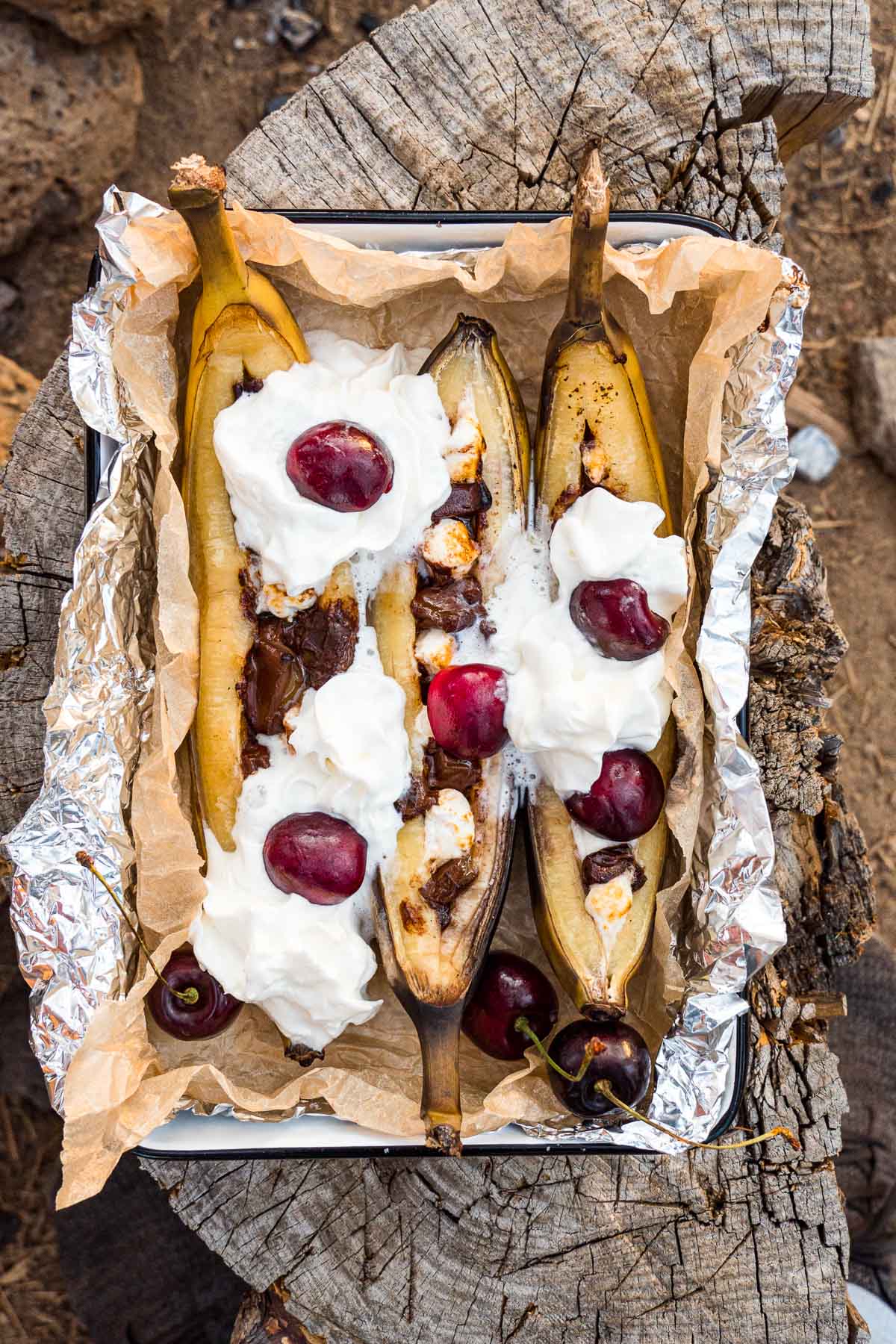Campfire banana splits topped with whipped cream in a serving dish on a log
