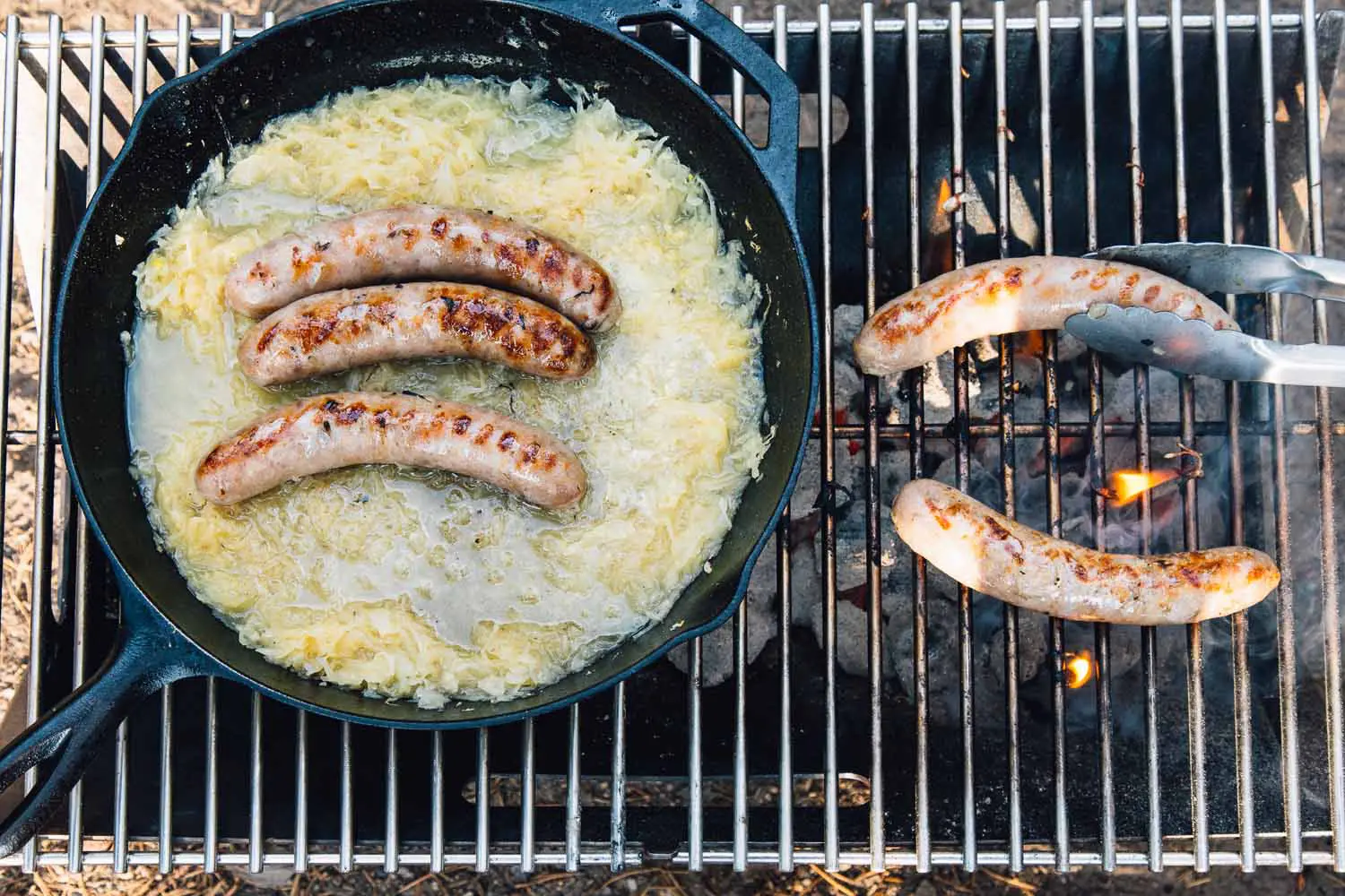 Overhead of a campfire with two beer brats on the grill and three beer brats simmering in beer and sauerkraut