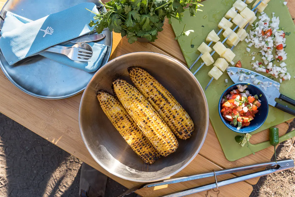 Three ears of grilled corn in a large bowl surrounded by other taco side dishes.