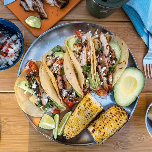 Four tacos arranged on a plate with grilled corn on the cob and hal an avocado