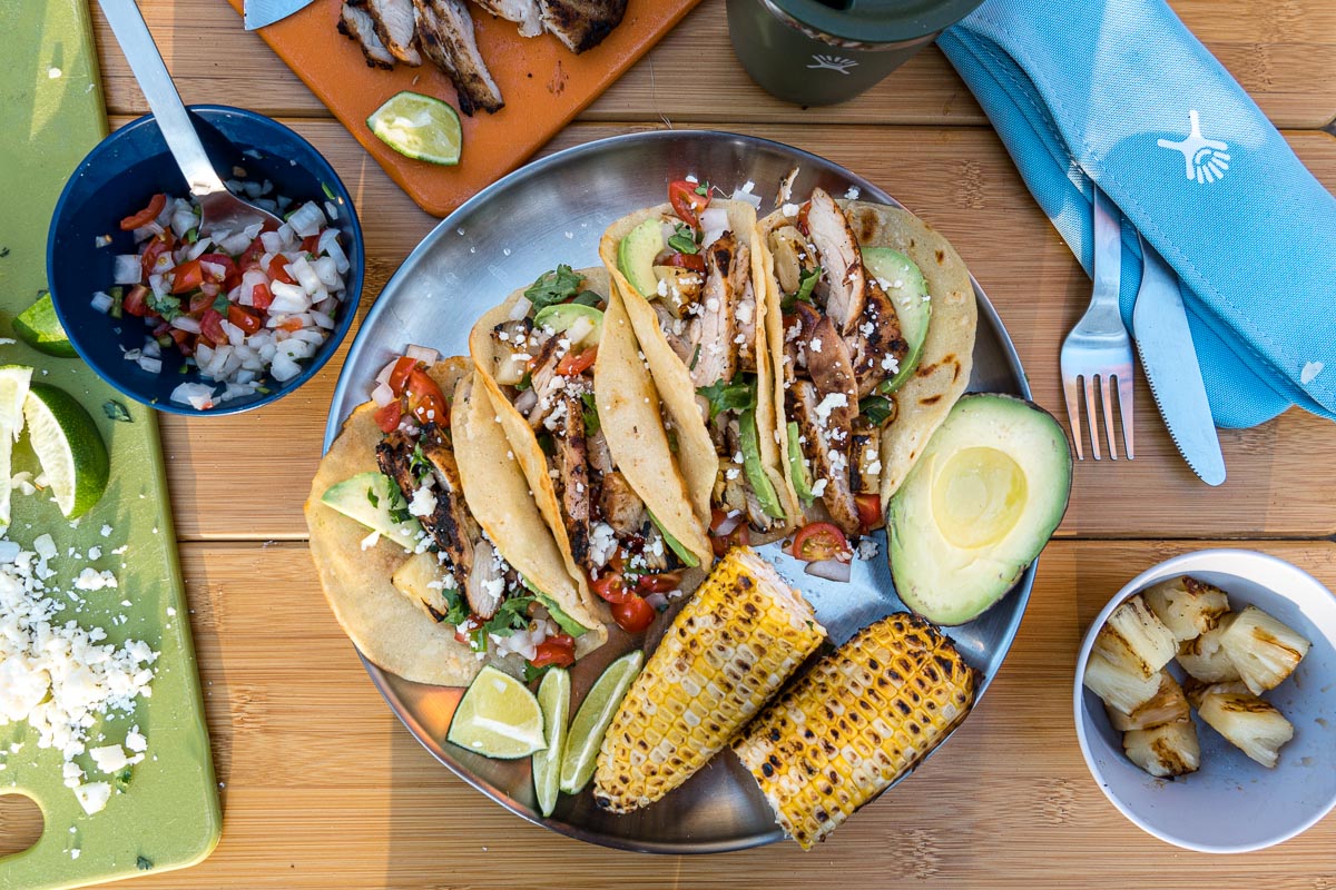 Four tacos arranged on a plate with grilled corn on the cob and hal an avocado
