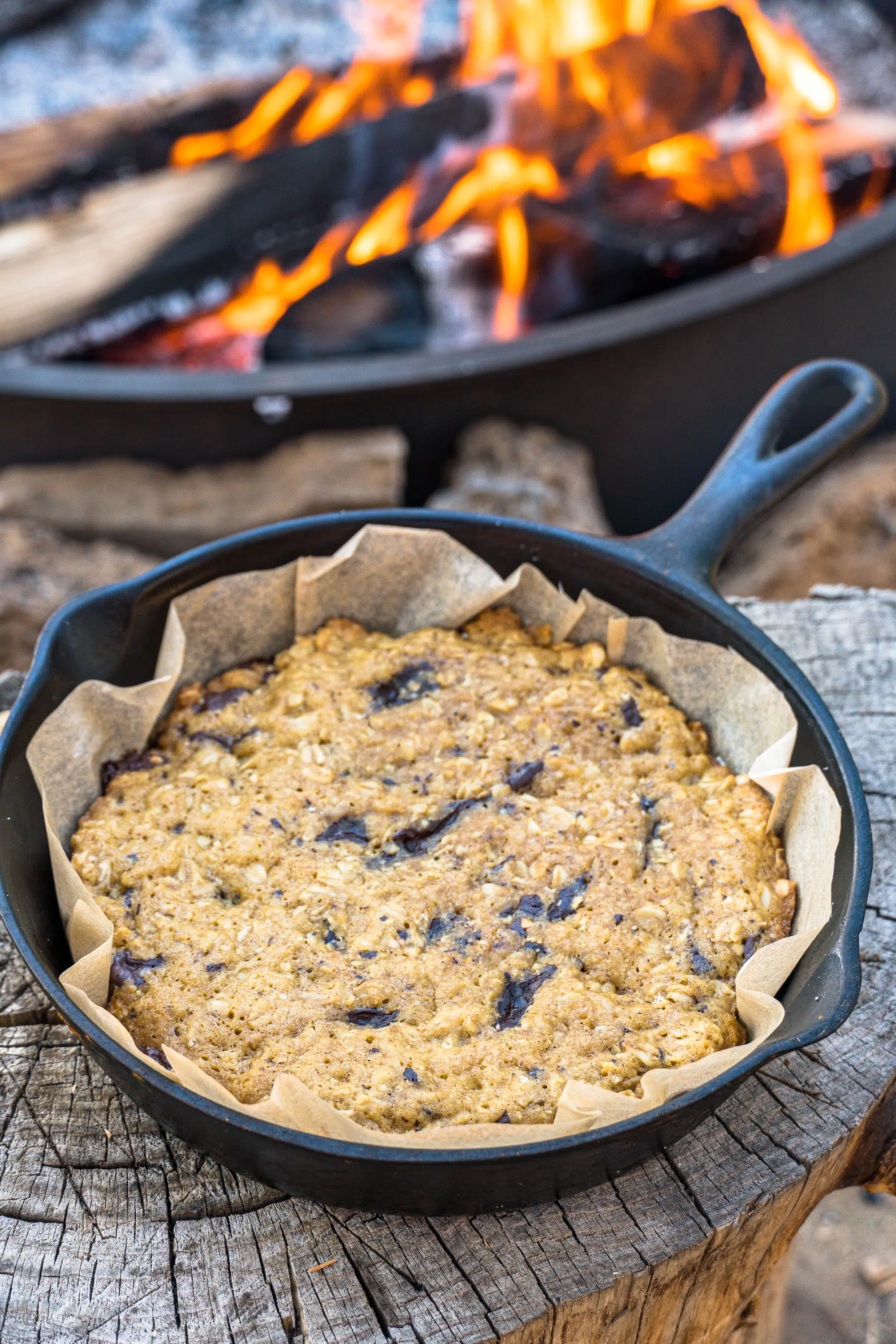 A chocolate chip cookie in a cast iron skillet set on a wood log.