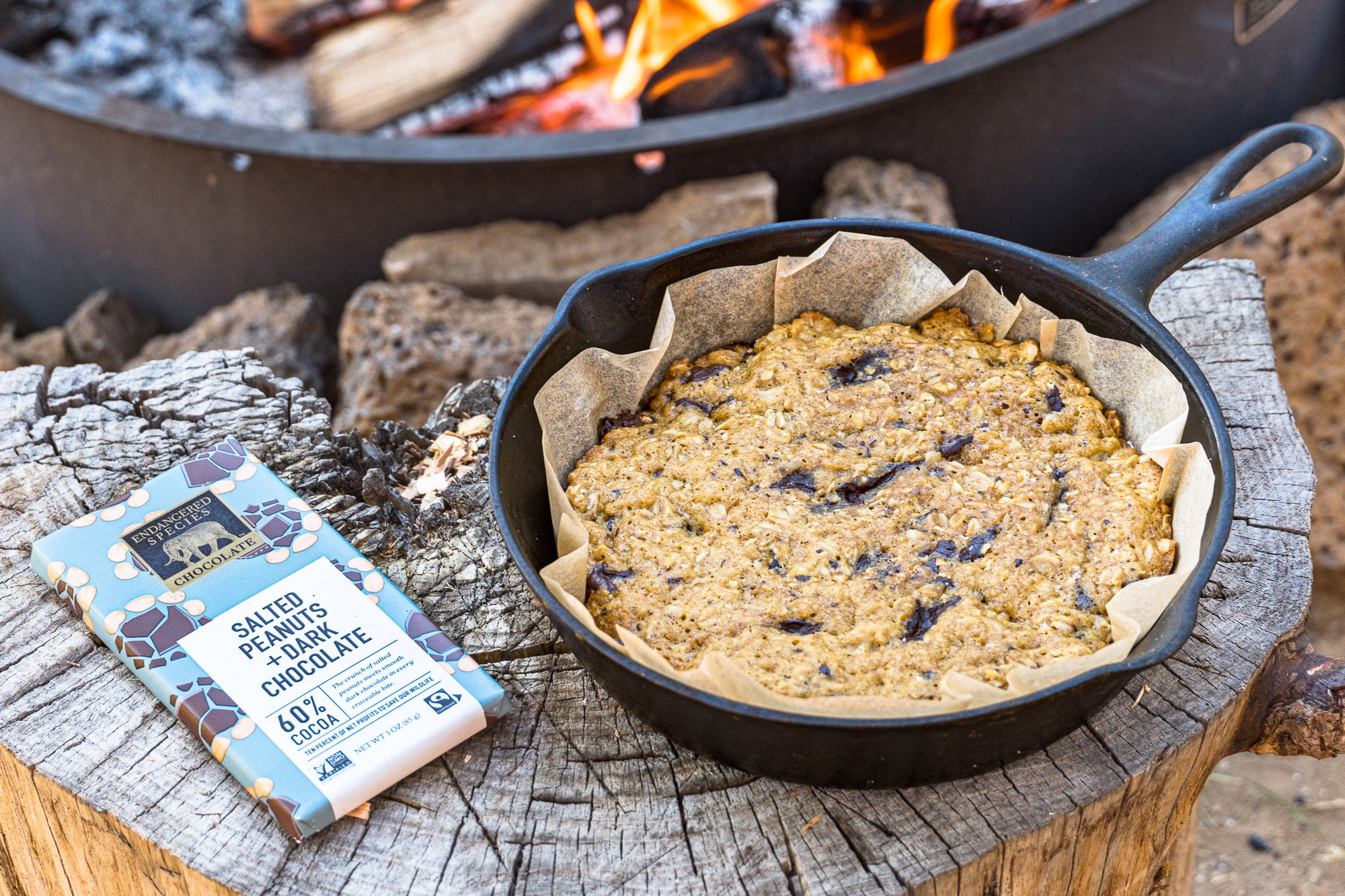 A chocolate chip cookie in a cast iron skillet set on a wood log.