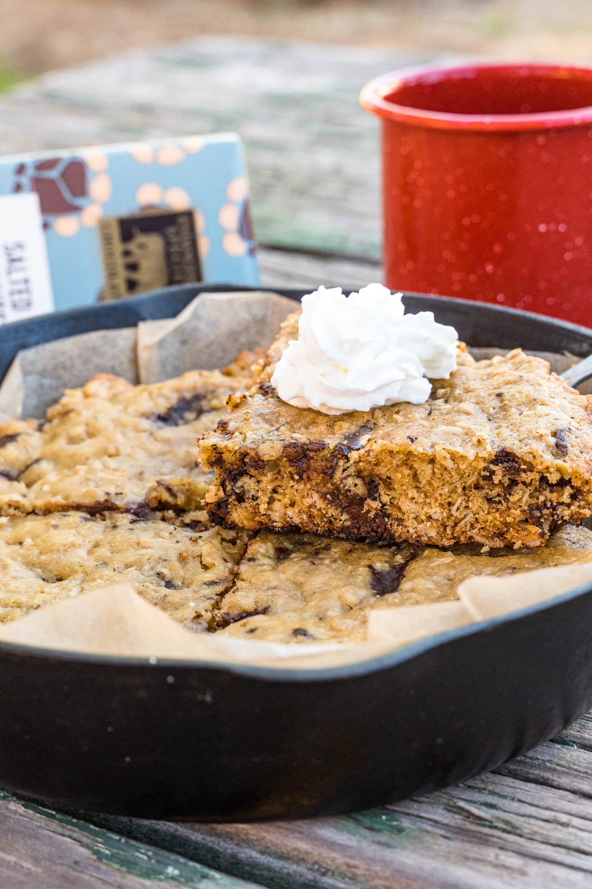 A chocolate chip cookie in a cast iron skillet. A slice has been cut out and is topped with whipped cream.