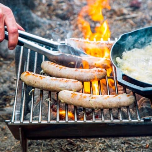 Grilling sausages over an open flame with onions sautéing in a pan nearby, capturing the essence of outdoor cooking.