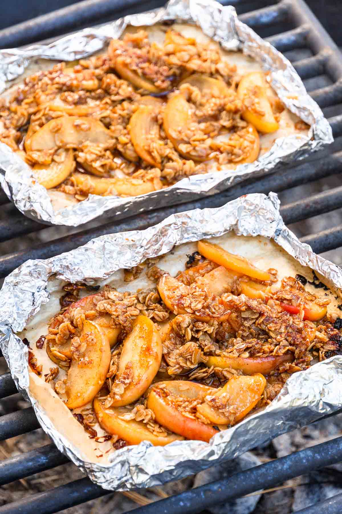 Two foil packets with baked sliced apples and a crumbly oat topping rest on a grill outdoors.