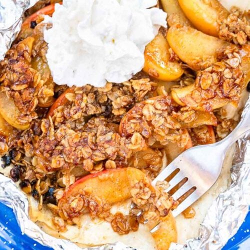 A close-up of a blue plate with foil holding apple crisp topped with oats and whipped cream, with a fork resting beside it.