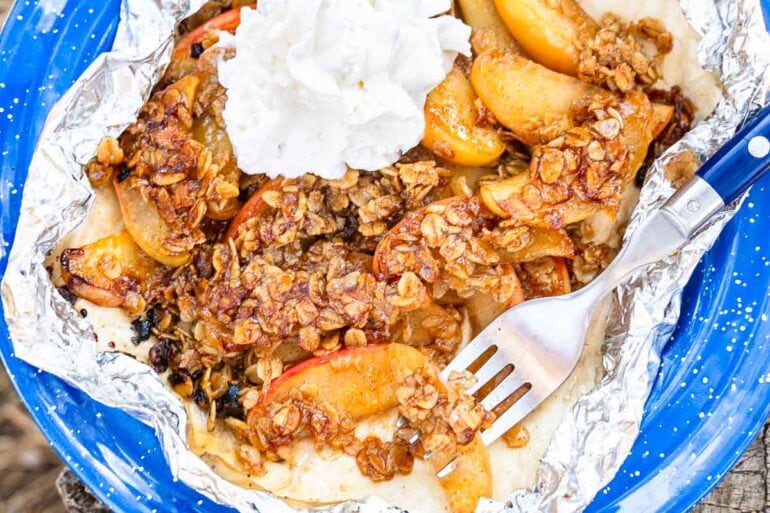 A close-up of a blue plate with foil holding apple crisp topped with oats and whipped cream, with a fork resting beside it.