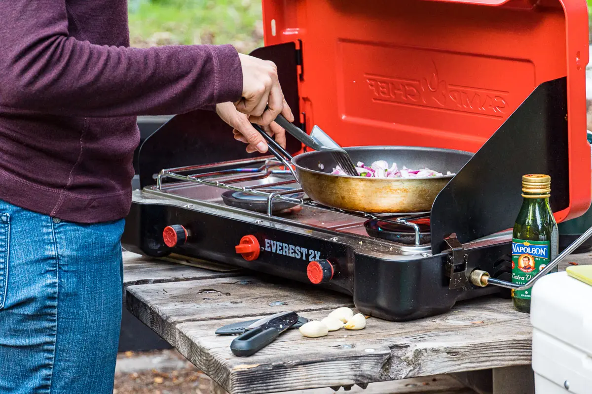 A camp stove sits on a picnic table. Megan's torso and hands are in frame as she sautes food in a skillet on the stovetop.