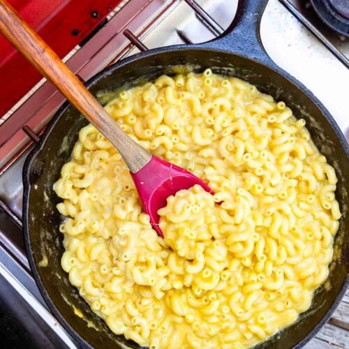 A cast iron skillet of macaroni and cheese sits on a stovetop, with a red and wooden spatula stirring the pasta.