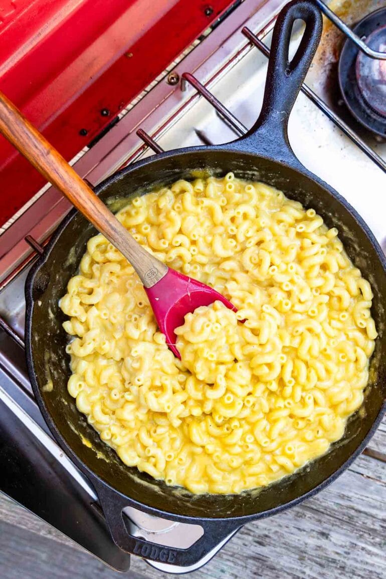 A cast iron skillet of macaroni and cheese sits on a stovetop, with a red and wooden spatula stirring the pasta.