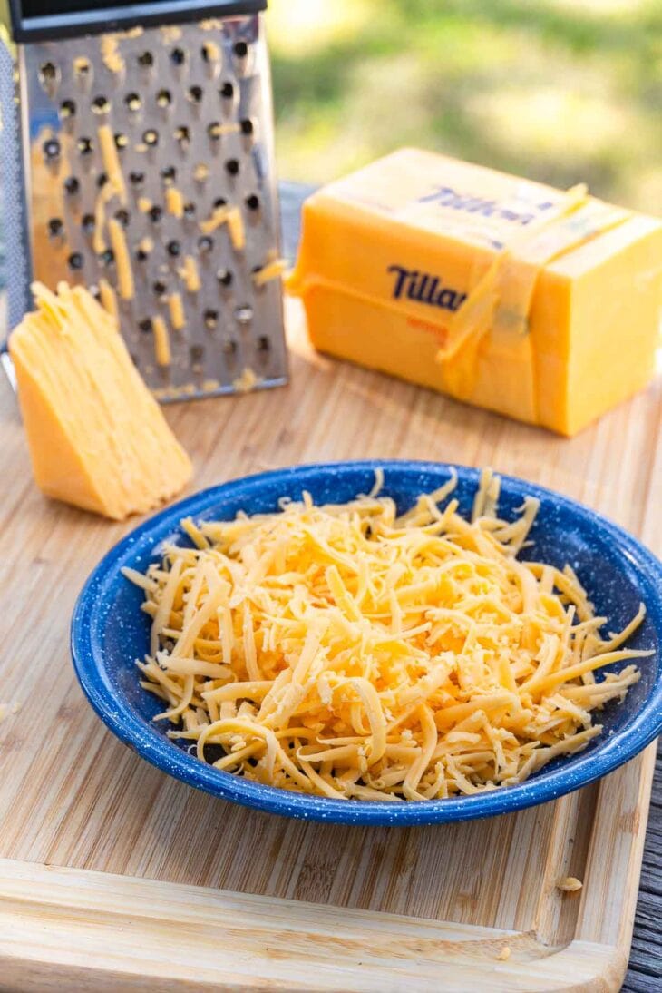 A blue speckled bowl of shredded cheddar sits on a wooden board beside a block of cheese, sliced cheese, and a metal grater.
