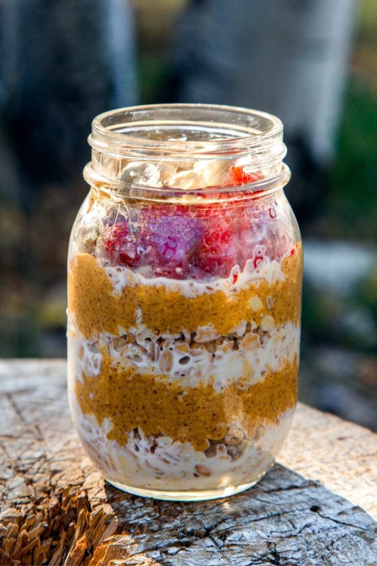 A mason jar with visible layers of oats, smooth nut butter, yogurt, and sliced strawberries rests on a wooden surface outdoors, with a blurred tree background.