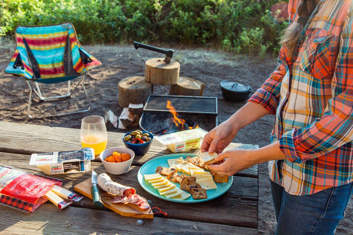 A woman with camping snacks on a picnic table with a campfire in the background.