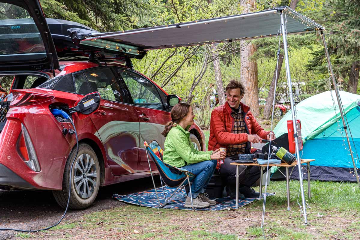 Megan and Michael sit next to a red car and are cooking on a camp stove.