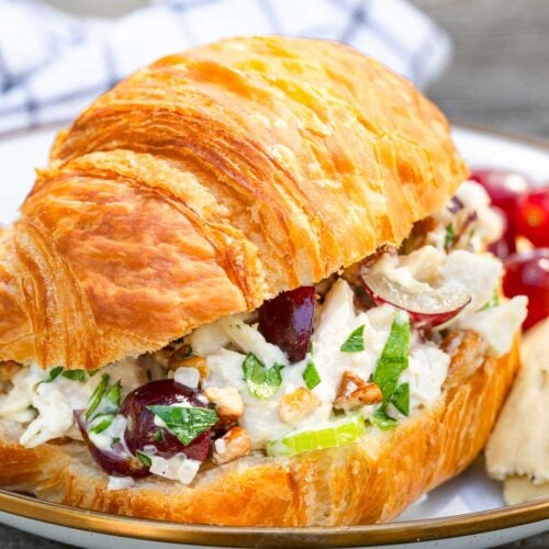 A croissant sandwich with chicken salad, grapes, celery, and pecans on a plate with grapes and a pastry in the background.