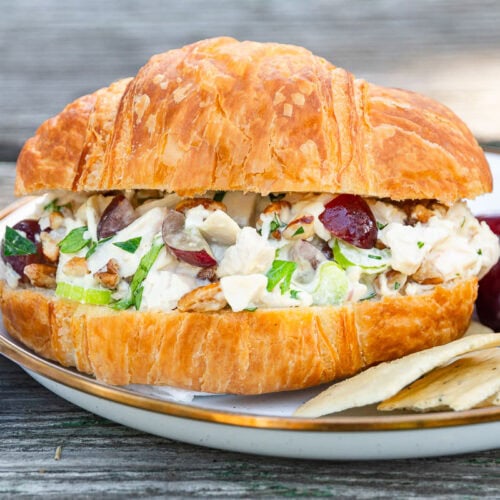 A croissant sandwich with chicken salad, grapes, and celery is on a plate with grapes and crackers beside a striped napkin.