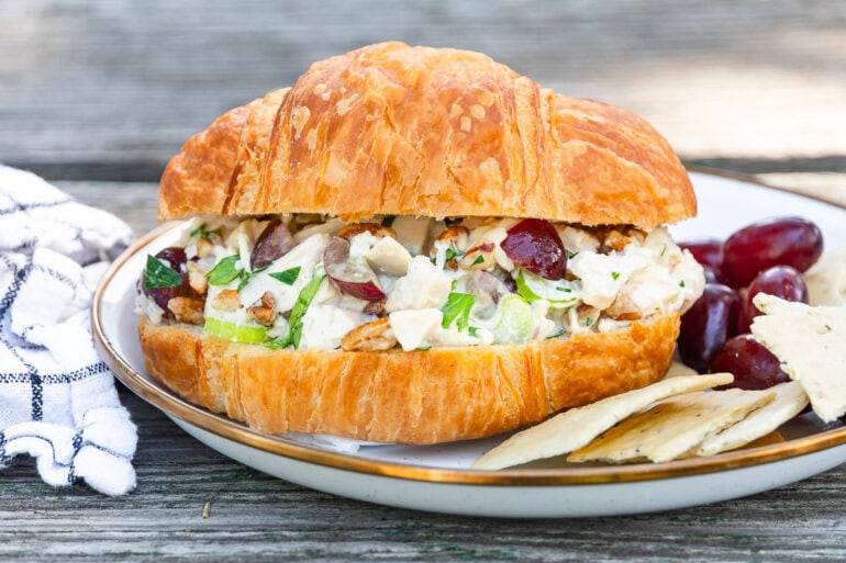 A croissant sandwich with chicken salad, grapes, and celery is on a plate with grapes and crackers beside a striped napkin.