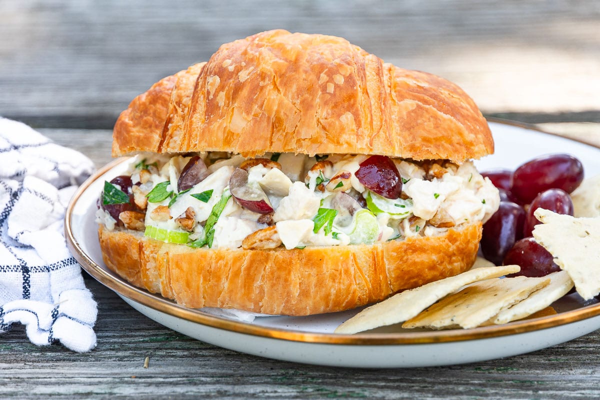 A croissant sandwich with chicken salad, grapes, and celery is on a plate with grapes and crackers beside a striped napkin.