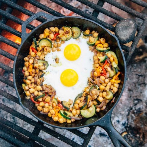 Chickpeas, vegetables, and two eggs in a skillet over a campfire.