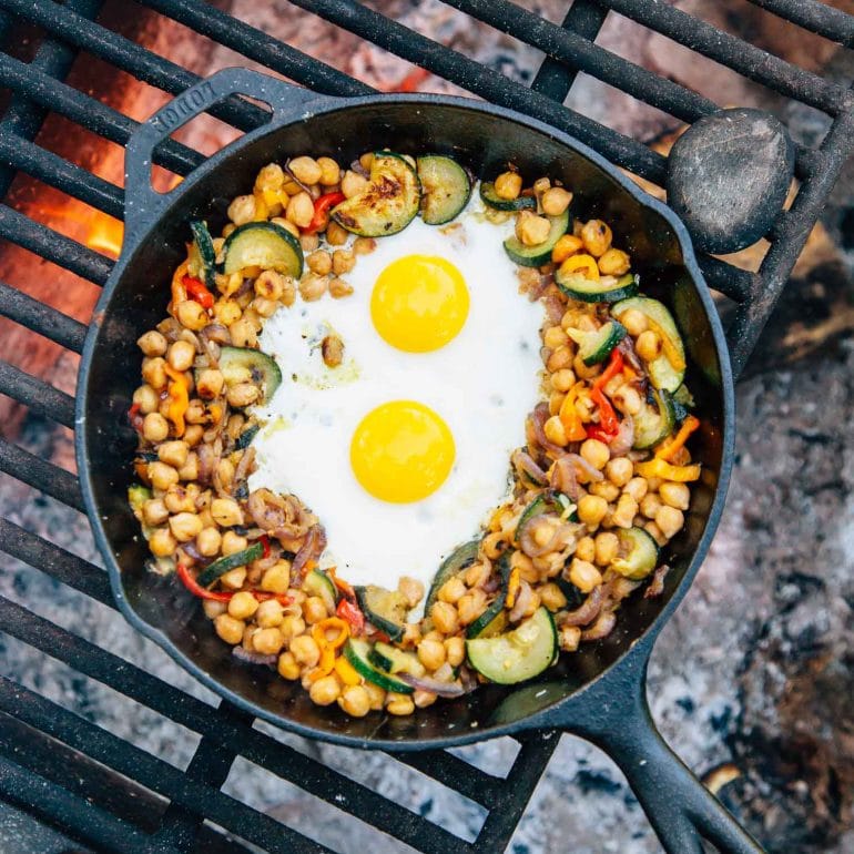 Chickpeas, vegetables, and two eggs in a skillet over a campfire.