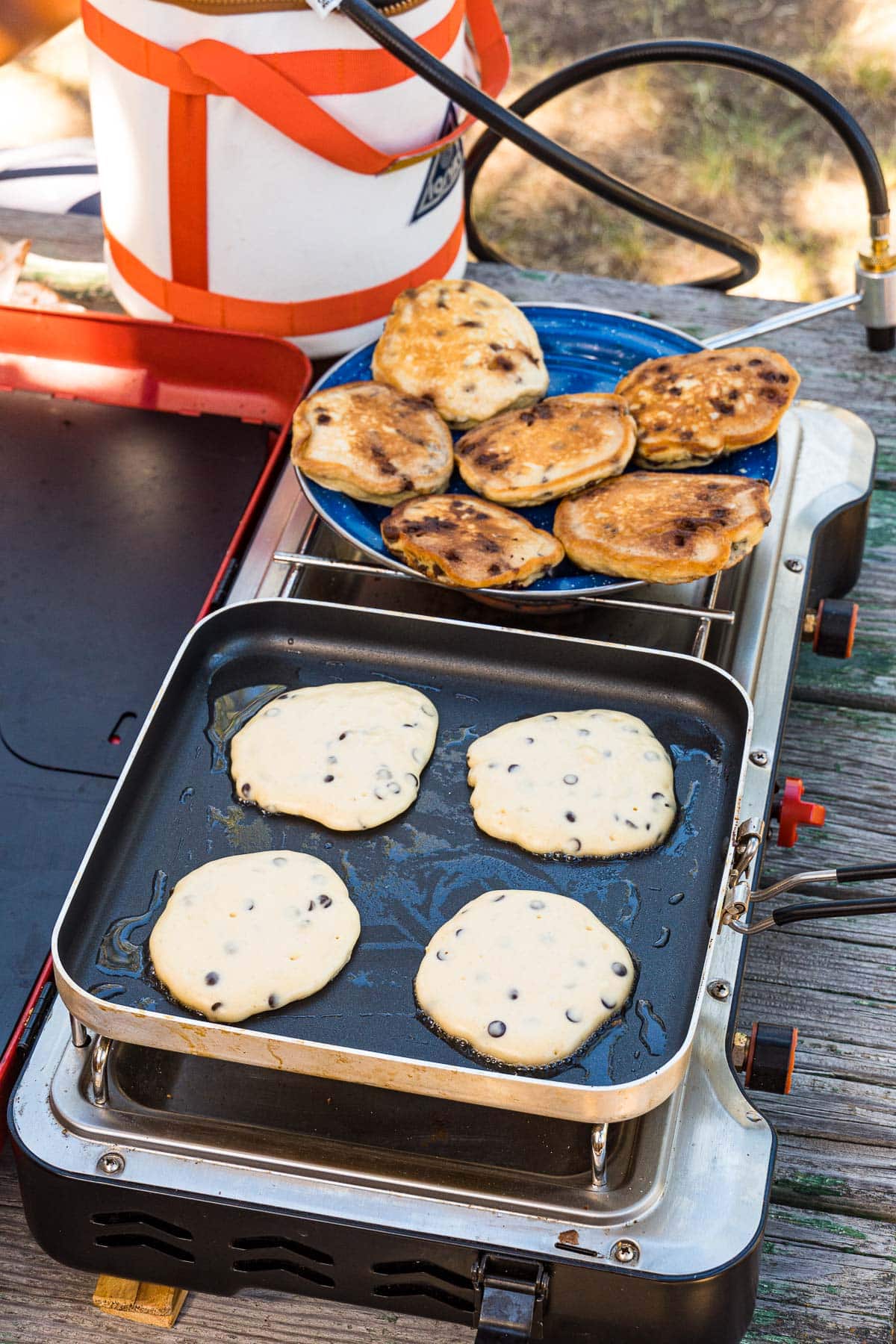 Pancakes in a skillet on a camp stove. To the side there is an enamel plate with cooked pancakes.