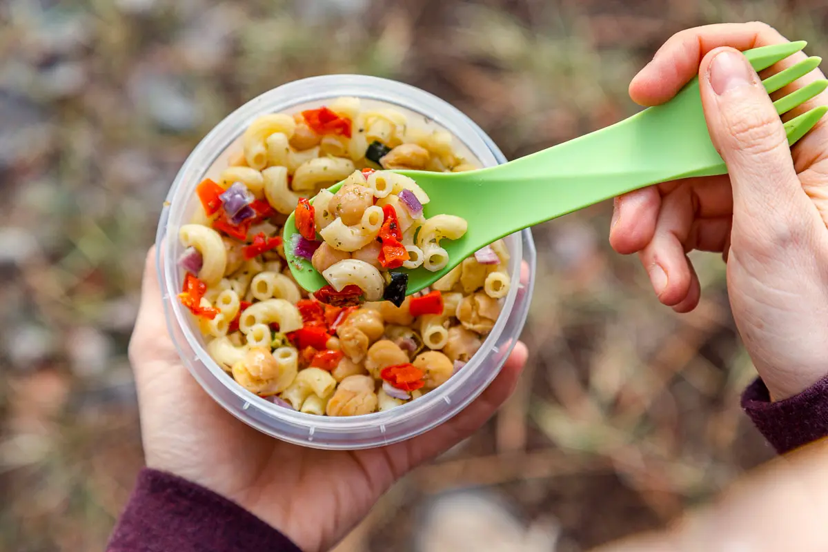 A person is outside with a portable pasta salad meal in a clear plastic container, paired with a green fork.