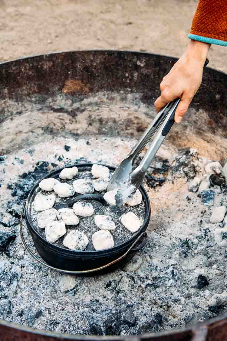 Woman adding coals to the top of a dutch oven in a campground fire pit.