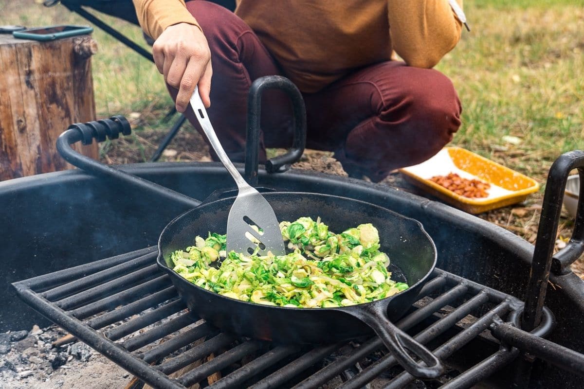 Megan crouched by a campfire cooking brussels sprouts in a cast iron skillet