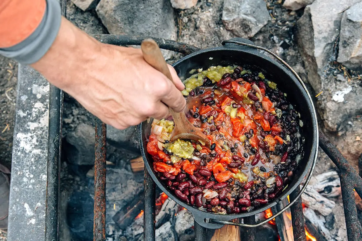 Michael stirring tomatoes, beans, and chilies in a Dutch oven over a campfire