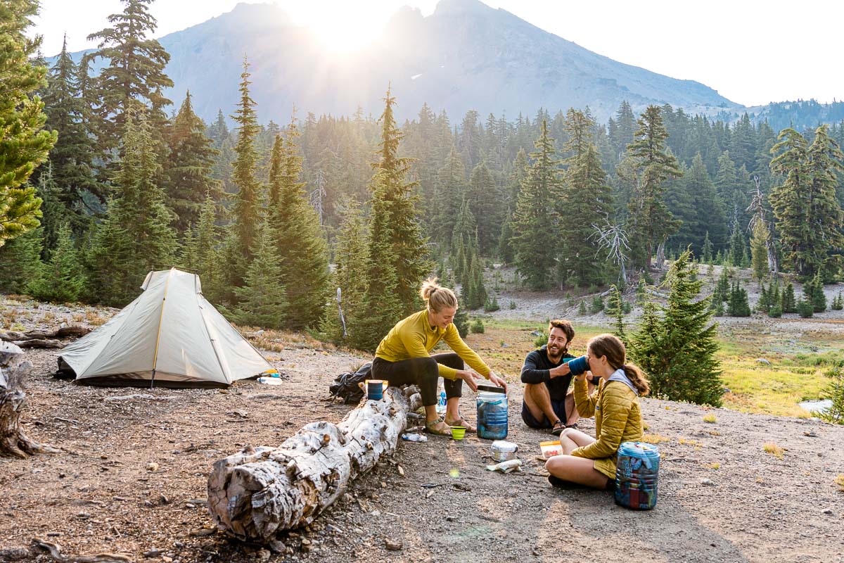 Three people cooking next to a backpacking tent with a mountain in the distance