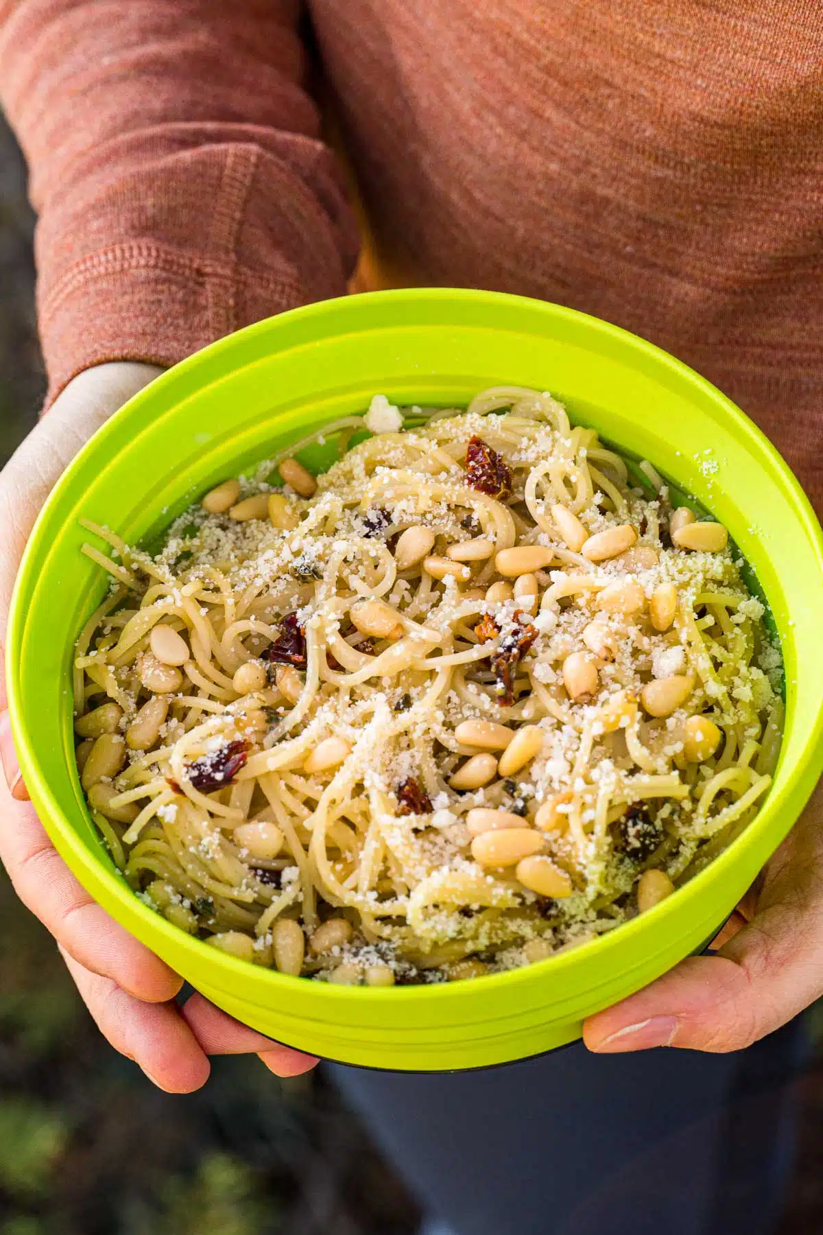 A person holding a bowl filled with pasta, pesto sauce, pine nuts, and sun dried tomatoes.