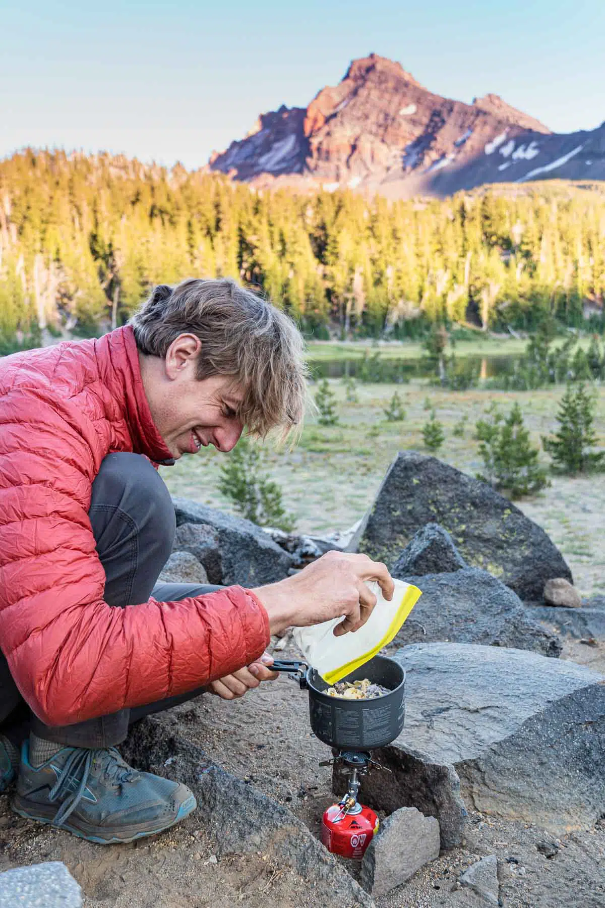 Michael andding food to a backpacking pot. There is a forest and mountain peak in the distance.