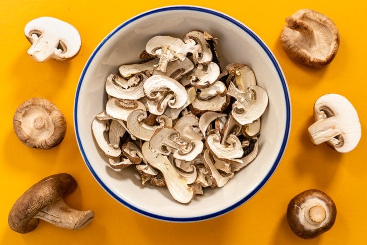 A bowl of sliced mushrooms surrounded by whole mushrooms on a yellow background.