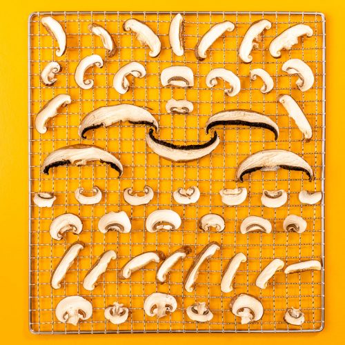 Sliced mushrooms artistically arranged on a wire rack to resemble a smiling face, set against a vibrant yellow background.