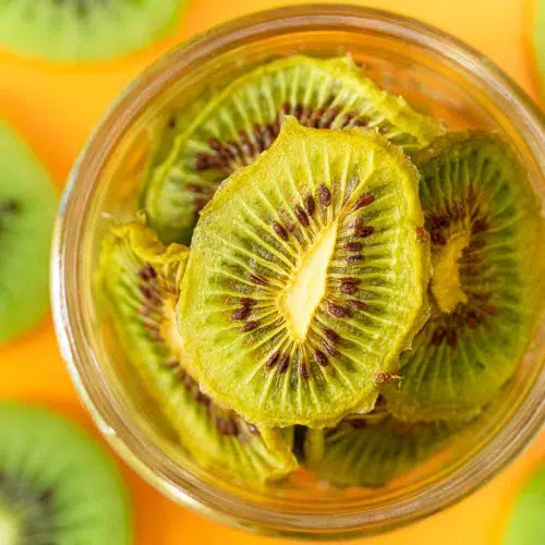 Dehydrated kiwis in a jar on a orange surface with sliced kiwis in the backgorund