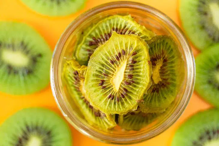 Dehydrated kiwis in a jar on a orange surface with sliced kiwis in the backgorund