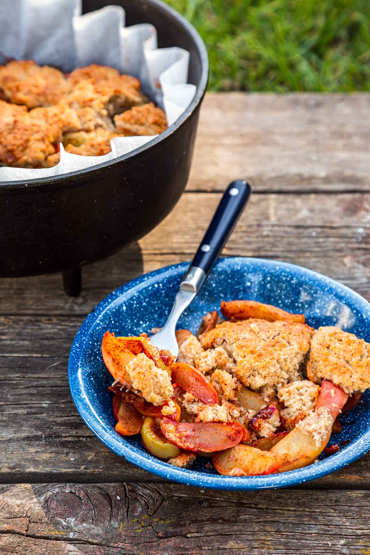 Apple cobbler in a blue bowl with a fork next to a Dutch oven