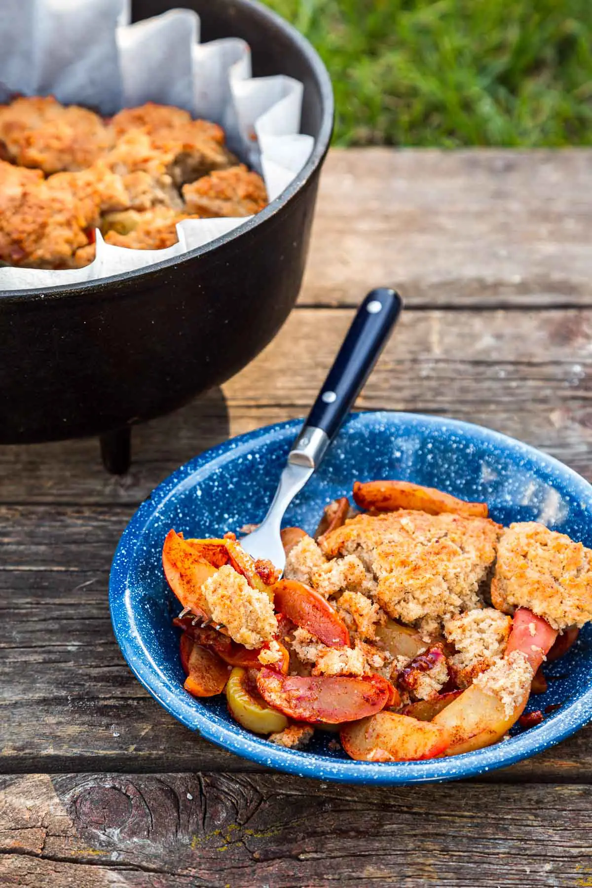 Apple cobbler in a blue bowl with a fork next to a Dutch oven