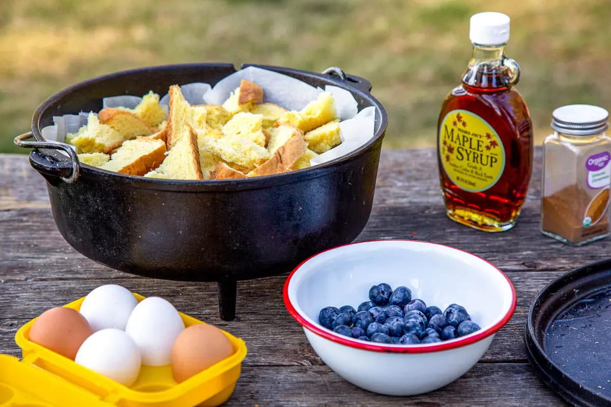 A dutch oven filled with torn bread, a bowl of blueberries, a yellow egg box with eggs, and a jug of maple syrup on a table.