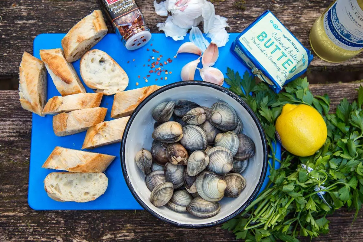 A table scene with a bowl of clams, sliced baguette, parsley, garlic cloves and a lemon on a cutting board
