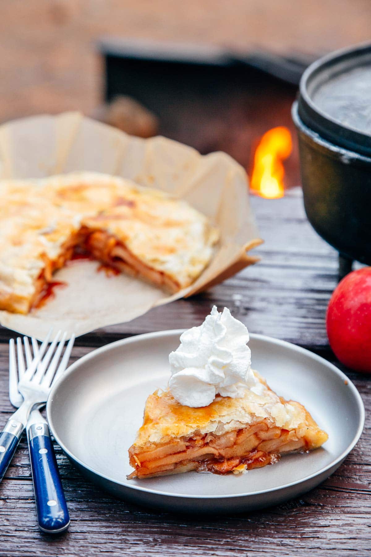 Slice of apple pie on a plate with a Dutch oven and campfire in the background