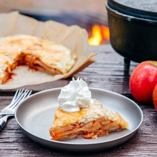 Slice of apple pie on a plate with a Dutch oven and campfire in the background