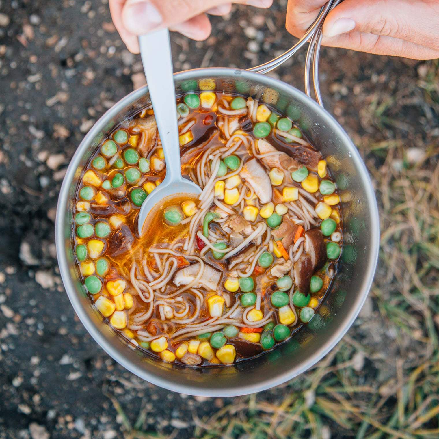 Ramen noodles, vegetables, and broth in a backpacking pot. A hand and spoon are reaching in.