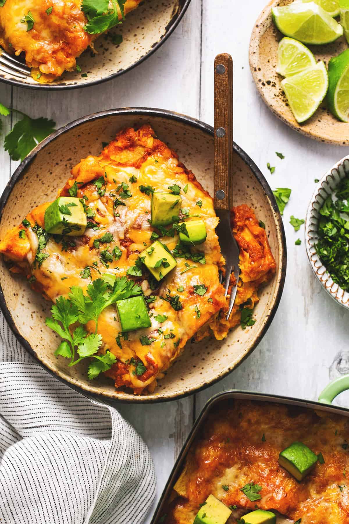 A freshly baked enchilada dish garnished with avocado, cilantro, and a lime wedge is displayed.